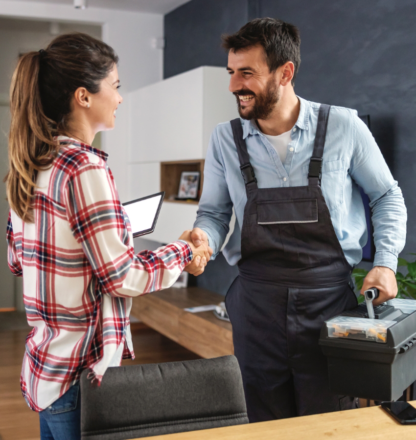 Handyman carrying a tool box shakes hands with a homeowner who is wearing a plaid shirt.